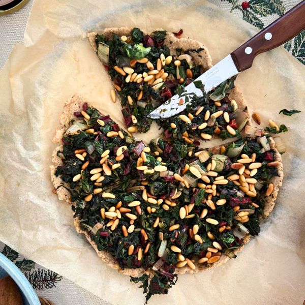 Esta tarta rústica tiene raíces profundas del mediterránea, evocando aquella icónica tourte de blettes de Niza (Francia), un pastel medieval elaborado desde el siglo XV con acelga, pasas, piñones y manzana, que combina lo salado y lo dulce y ha acompañado la mesa de muchas generaciones. En esta versión ayurvédica, la masa se prepara con harina integral y de centeno, lo que la hace más nutritiva y satisfactoria, mientras que el relleno de acelga con especias digestivas como hinojo, cardamomo y canela le da un carácter aromático y equilibrado. Los piñones tostados añadidos al final aportan crocancia y un toque elegante que transforma la sencillez en algo especial. Es un plato que conecta lo rústico con lo refinado: puede servirse tibio como un desayuno salado diferente, como parte de una comida principal acompañado de una sopa de legumbres y verduras que construyen, o bien con un poco de queso fresco y una guarnición de verduras para disfrutarlo como una comida completa. Es ideal en invierno, otoño y primavera, cuando el cuerpo agradece preparaciones tibias, especiadas y nutritivas que reconfortan sin dejar pesadez.