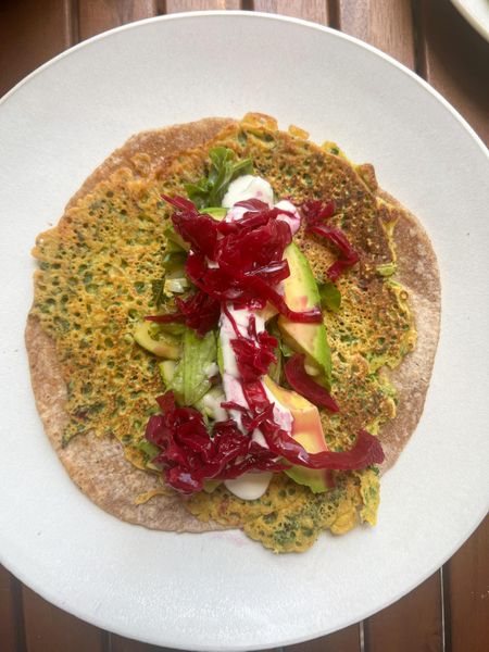 Había visto un trend en redes sociales donde preparaban tipo omelettes pegados a un pan; esta es mi versión ayurvédica, inspirada en ese concepto pero usando harina de garbanzo. El resultado es un wrap de falafel exprés nutritivo y muy fácil de hacer, que además resulta un éxito con los niños. La harina de garbanzo combinada con hierbas y especias se cocina como una fina tortilla que se adhiere al chapati y forma una base rica en proteína vegetal y de sabor profundo. El relleno se puede personalizar, pero recomiendo mantenerlo equilibrado: aguacate, calabacín salteado y un toque de chucrut o sauerkraut para favorecer la digestión. La salsa de yogur con tahini lo convierte en un wrap completo, cremoso y saciante. Es ideal en invierno, primavera e inicios del verano como comida ligera, rápida y nutritiva, perfecta para el mediodía o como desayuno más sustancioso. 