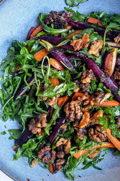 La mayoría de las ensaladas que se sirven en Navidad… son una bomba para la digestión. Mucha verdura cruda, fría, pesada… a veces mezclada con frutas y lácteos, y encima se sirve junto a un plato principal que ya de por sí suele ser muy pesado. Esa combinación casi siempre termina en inflamación, gases y la clásica sensación de “me pasé”. Por eso preparé esta versión Ayurvédica: festiva, hermosa en la mesa, pero suave para tu cuerpo. Aquí usamos arugula en vez de lechuga cruda; su sabor ligeramente pungente y su cualidad más cálida la hacen mucho más digestiva. Las zanahorias se hornean hasta quedar dulces, tiernas y con un toque caramelizado que aporta estabilidad y calidez, perfectas para el otoño e invierno, cuando el cuerpo necesita más tierra y fuego suave. El toque final lo dan las nueces caramelizadas de manera natural, sin azúcar refinada, que aportan crunch y un dulzor ligero sin saturar el sistema digestivo. El resultado es una guarnición colorida y nutritiva que acompaña tu comida en vez de competir con ella. Una ensalada tibia, festiva y equilibrada, perfecta para el mediodía o una cena ligera… hecha para que disfrutes la temporada sin sufrir después.