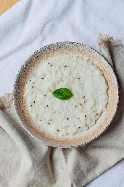 Hay comidas que no están hechas para impresionar, sino para acompañar al cuerpo cuando más lo necesita. Este arroz con yogur diluido es una de ellas. En muchas tradiciones, esta preparación (o alguna muy similar) ha sido el primer alimento que se introduce después de una enfermedad digestiva. Cuando has pasado por un virus estomacal, cuando el apetito desaparece… y poco a poco regresa, este tipo de comida es el puente entre el ayuno y volver a comer con normalidad. Porque no exige. Sostiene. El arroz, bien cocido y ligeramente caldoso, es fácil de digerir y le da al cuerpo una fuente de energía estable sin sobrecargar el sistema. El yogur, en lugar de usarse denso y pesado, se diluye hasta convertirse en algo ligero, casi como una bebida. Esto cambia completamente su efecto en el cuerpo. En Ayurveda, esta forma más ligera se acerca a takra (buttermilk), que estimula la digestión sin generar pesadez ni mucosidad, como puede hacerlo el yogur espeso. Además, cuando el sistema digestivo ha pasado por una infección, la microbiota intestinal suele verse afectada. Aquí es donde esta preparación puede apoyar suavemente: el yogur diluido aporta bacterias beneficiosas en una forma más fácil de tolerar, mientras que la textura ligera del plato permite que el intestino reciba nutrición sin sentirse invadido. No se trata de “reparar” todo de golpe, sino de crear el entorno adecuado para que el cuerpo vuelva a organizarse. Y luego está el toque final: el tadka de semillas. El eneldo (o el comino) no está ahí solo por sabor. Su función es sutil pero clave: ayuda a reducir gases, favorece la digestión y evita que esta preparación, aunque suave, se vuelva pesada o estancada en el intestino. Es ese pequeño gesto el que transforma el plato de “blando” a verdaderamente digestivo.