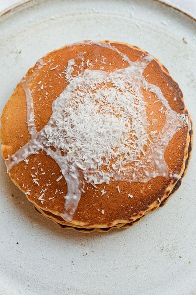 Ya saben que los pancakes son algo muy presente en nuestra cocina y cada temporada aparece una versión nueva. Esta la hice por primera vez la semana pasada y estoy segura de que se va a quedar. Rayyan los devoró, que ya es la mejor señal. El coco aparece mucho en nuestra cocina durante el verano, y no es casualidad. Tiene una cualidad naturalmente refrescante y al mismo tiempo muy nutritiva, hidrata, aporta grasas de buena calidad que el cuerpo asimila con facilidad, y ayuda a calmar el exceso de calor interno sin dejar esa sensación de pesadez que otros alimentos más densos pueden generar. Es uno de esos ingredientes que en esta época simplemente tiene sentido. Estos pancakes son ligeros pero satisfactorios. La leche de coco entera les da una textura suave y húmeda, y el coco rallado aporta un punto de textura muy agradable. La espelta o el trigo sarraceno los hacen más digestivos que la harina blanca convencional, y el hinojo en la masa ayuda a que todo se digiera con facilidad, un detalle pequeño pero que marca la diferencia. El reposo de cinco a diez minutos antes de cocinarlos también importa, así que no lo saltes. Sírvelos con duraznos o albaricoques salteados en ghee, una cucharada de yogur de coco, o simplemente un chorrito de miel al final. Un desayuno de verano que se prepara en veinte minutos y gusta a grandes y chicos.